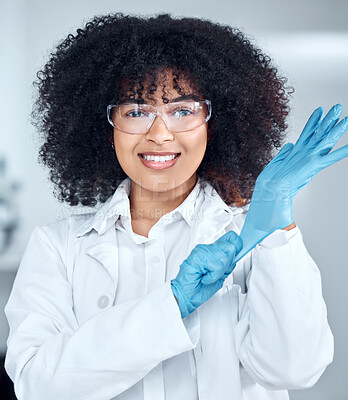 Buy stock photo Portrait of young african american female scientist with afro hair wearing a labcoat and goggles while putting on gloves in the laboratory. A mixed race female scientist getting ready to conduct an experiment 