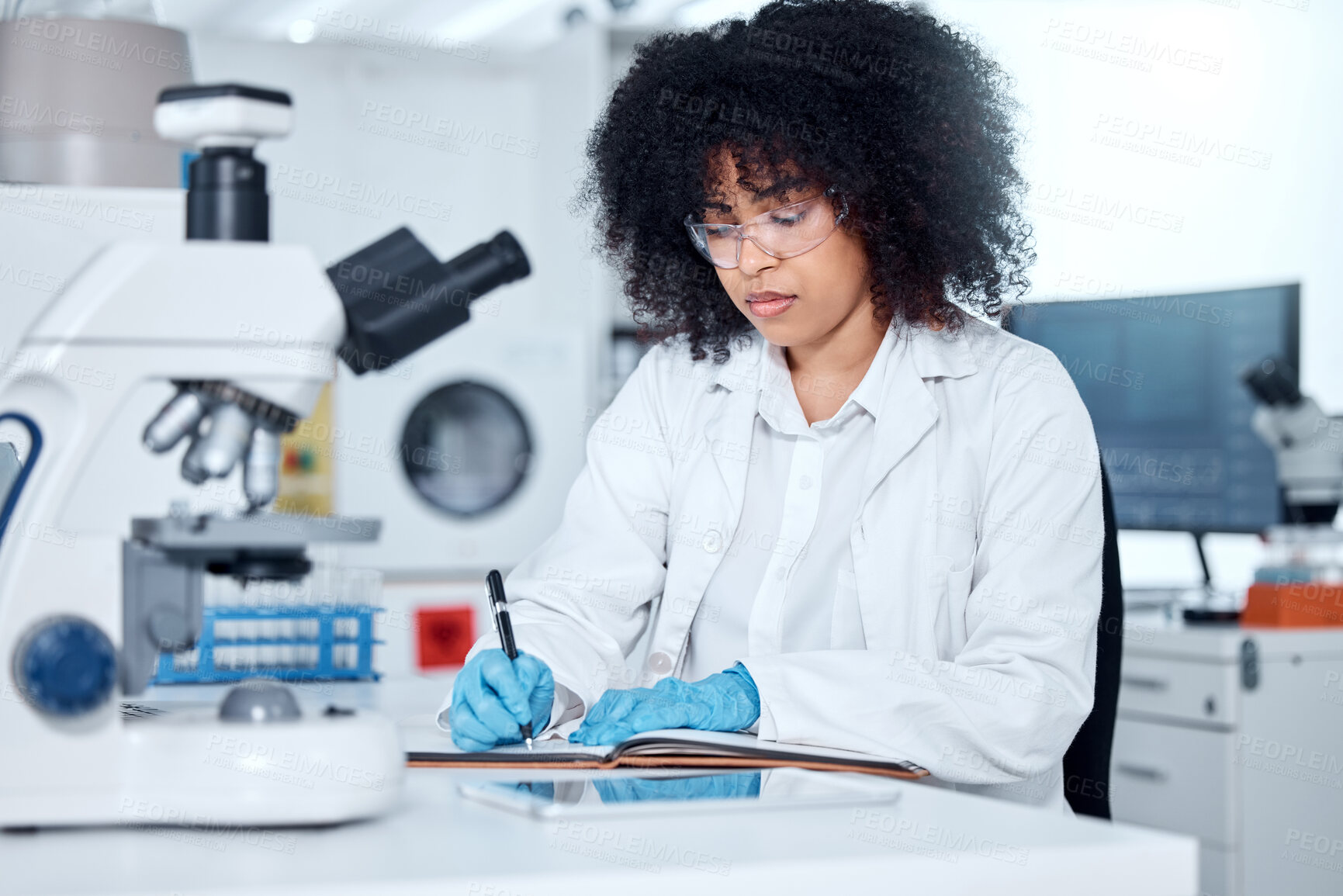 Buy stock photo One mixed race scientist with curly hair wearing safety equipment writing notes and analysing medical test samples with microscope in lab. Woman doing forensic research and experiments to develop cure