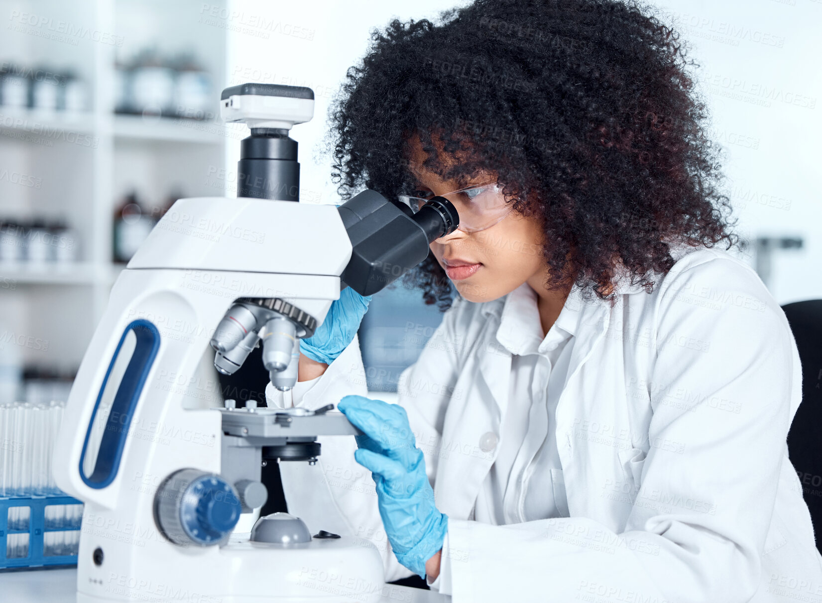 Buy stock photo Young african american woman wearing a labcoat and goggles looking at medical samples on a microscope in her lab. A mixed race female scientist wearing goggles and gloves conducting research
