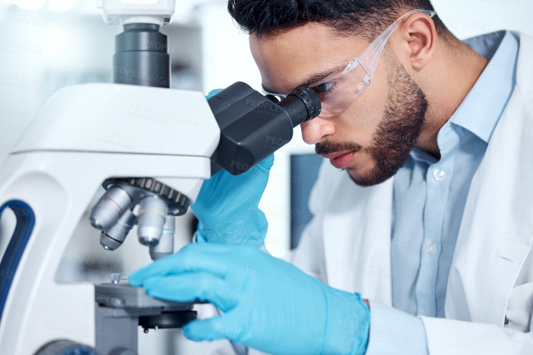 Buy stock photo One handsome young mixed race man with a dark beard wearing gloves and a labcoat and looking at medical samples on a microscope in her lab. A male scientist of indian ethnicity wearing safety goggles