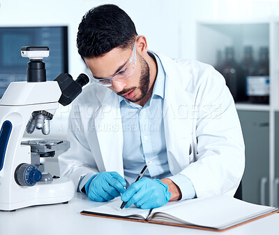 Serious young mixed race male scientist wearing glasses and gloves while writing in a notebook sitting at a desk alone working in a lab. One hispanic lab worker wearing goggles and taking notes