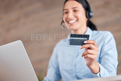 Happy young mixed race call center agent holding a credit card in her hand and working on a laptop while wearing a headset and answering calls in an office at work