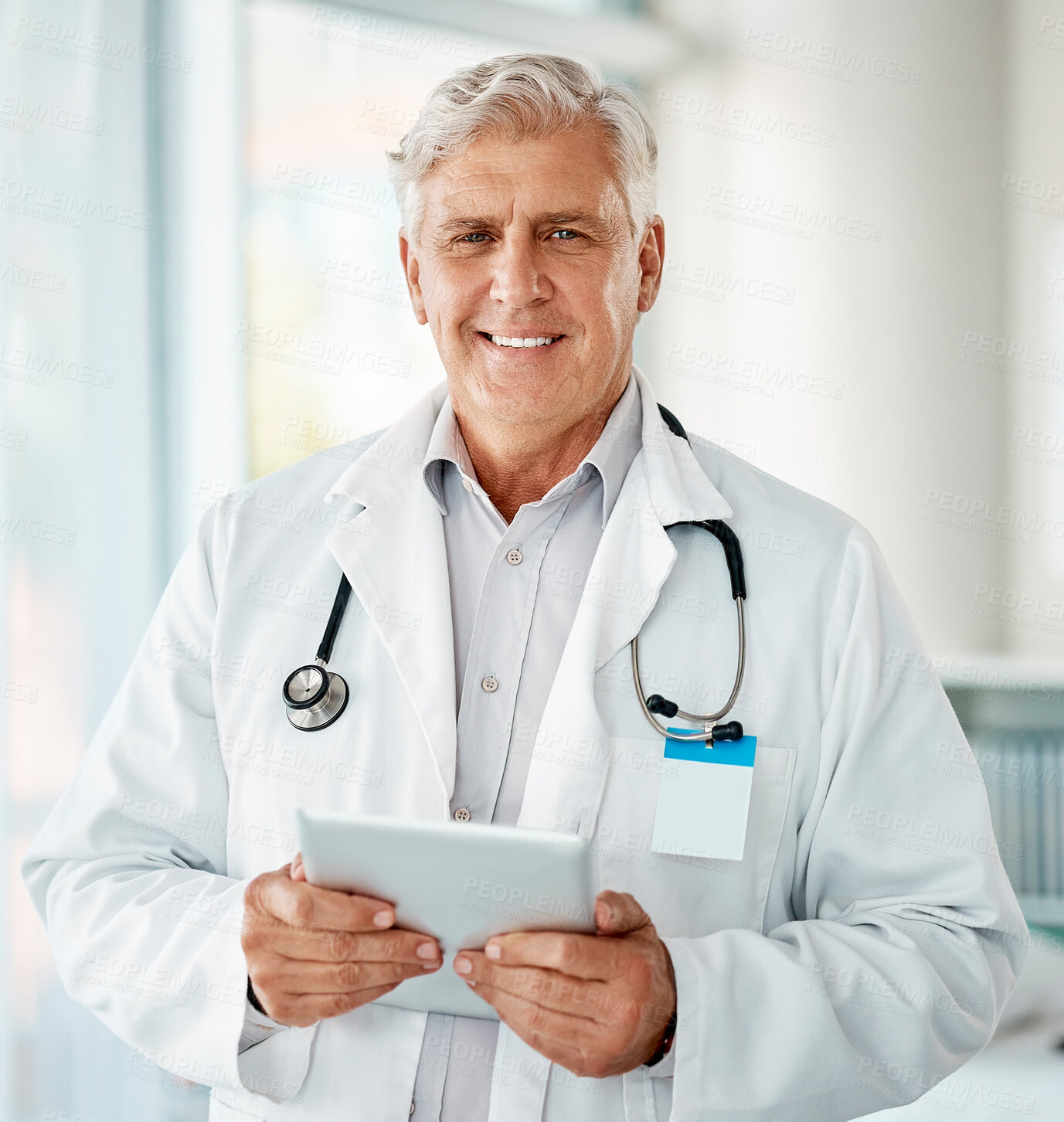 Buy stock photo Mature caucasian male expert doctor smiling while using a digital tablet working at a hospital alone. One senior man wearing a labcoat and working on a digital tablet standing in an office at a clinic