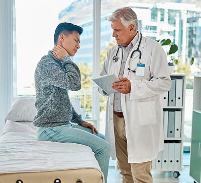 Buy stock photo Mature medical doctor standing and using a digital tablet while checking on his injured asian patient in a clinic. Young mixed race man suffering from a painful neck in a hospital. Tension and sprain