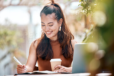Young happy mixed race businesswoman writing in a notebook drinking coffee and working on a laptop sitting outside at a cafe. Hispanic female student smiling while studying at a restaurant