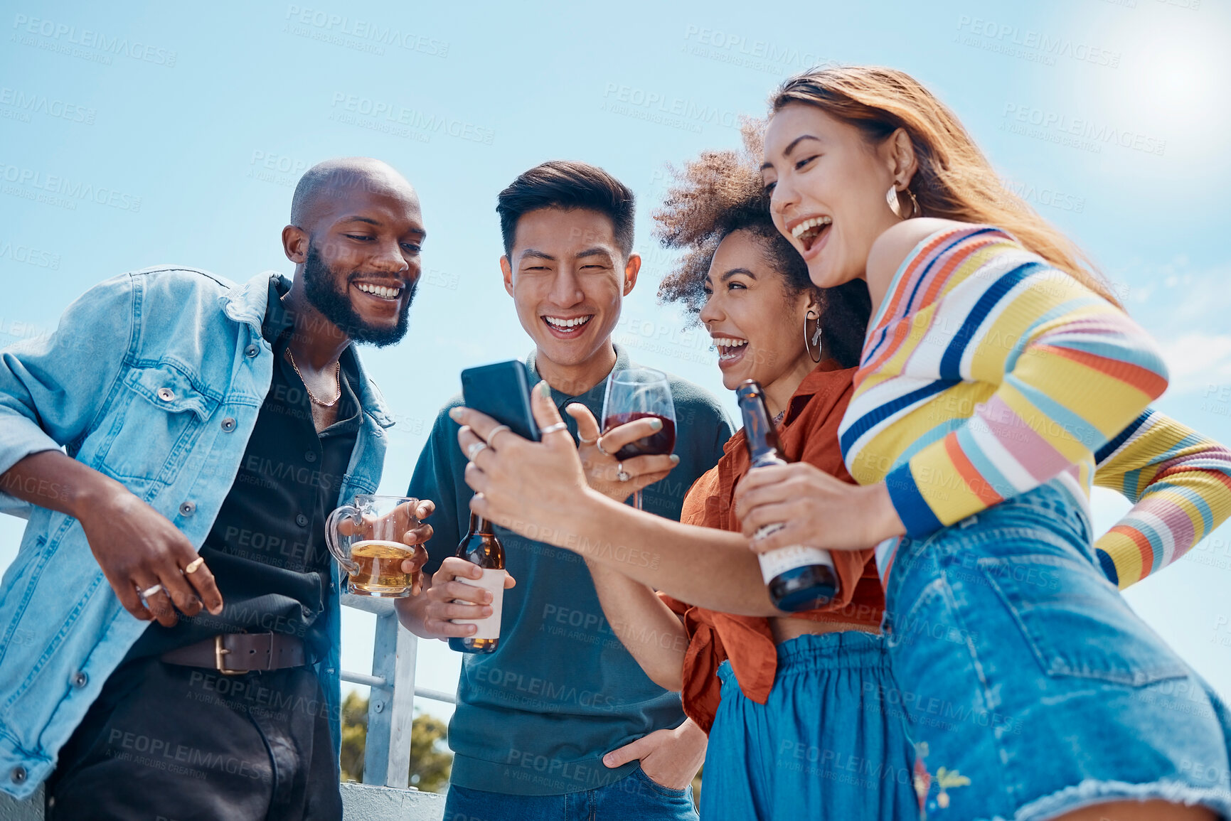 Buy stock photo A group of diverse young cheerful friends standing outside together and using a cellphone for selfies and social media. Smiling men and women being social and celebrating on a rooftop weekend party 