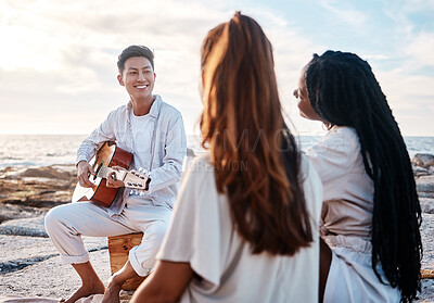 Buy stock photo A young man at the beach playing his guitar with his two female friends. Diverse group of friends listening music on a sunny day