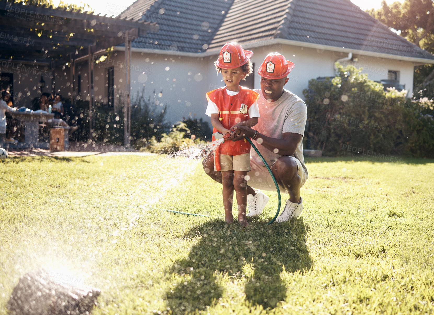 Buy stock photo Father playing with his son outside. Little boy dressed as firemen. African American boy playing outside on a sunny day. Young male playing with a hosepipe in the garden. Dad and son spraying water from a hosepipe.