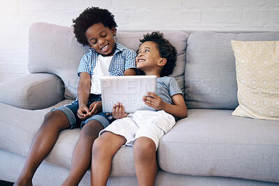 Two cute little african american boys sitting comfortably on a sofa using a wireless tablet while browsing and smiling. Black sibling brothers with cool afro hair style having fun at home