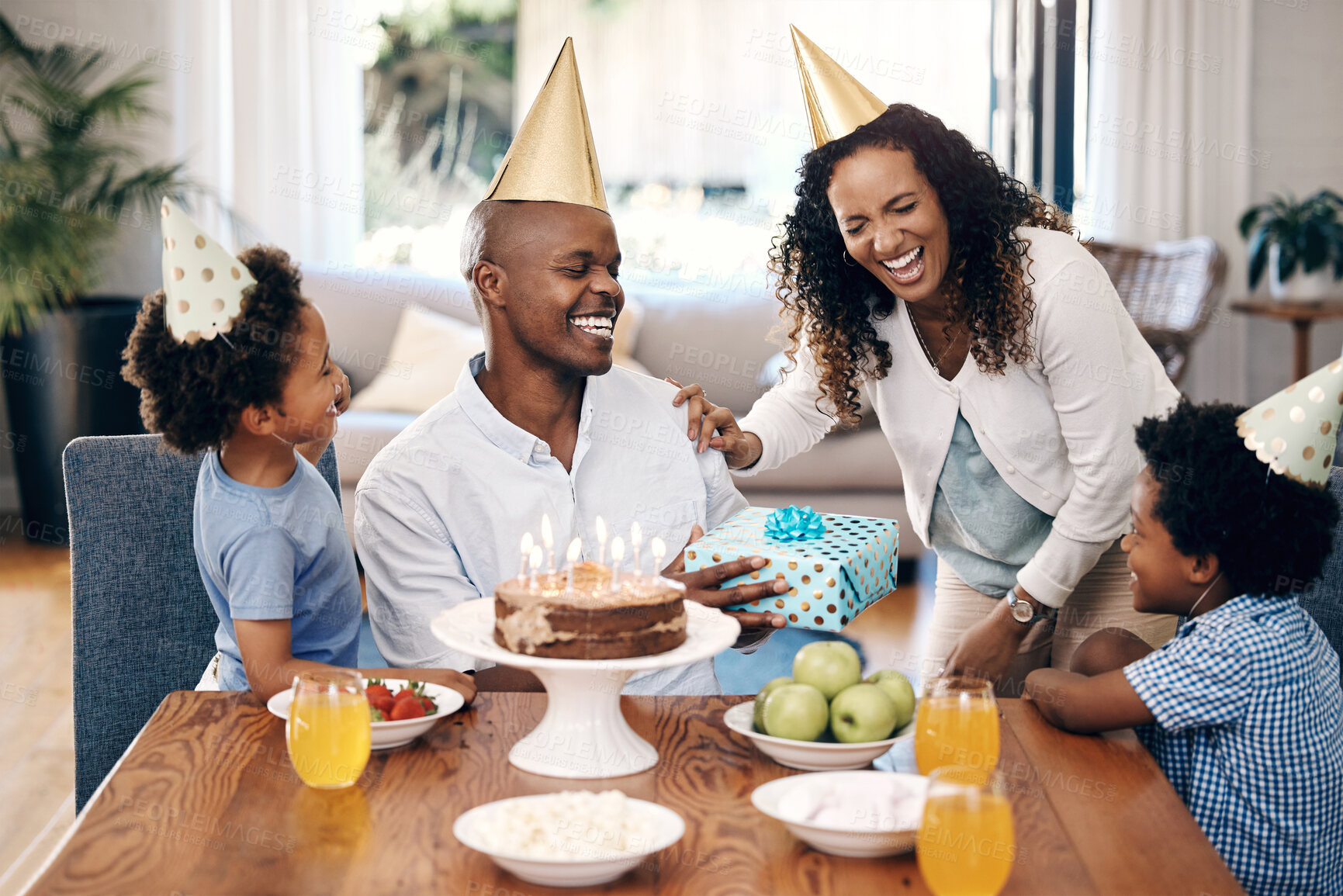 Buy stock photo Happy african american family celebrating a birthday at home. Cheerful parents and their two kids laughing while wearing party hats. Boy receiving present from family on his birthday