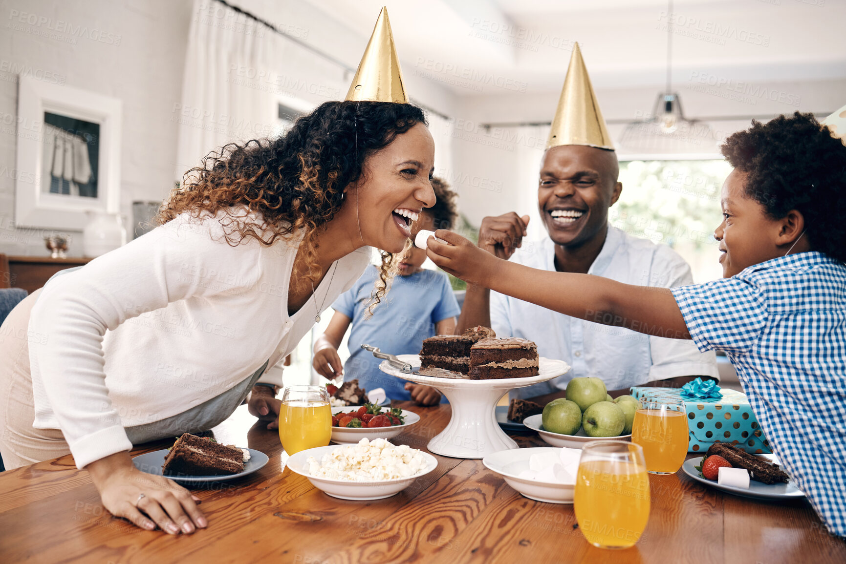 Buy stock photo Little boy feeding his cheerful mother a marshmallow while celebrating wearing party hats. African american family celebrating the birthday of their children eating sweet snacks at a party at home
