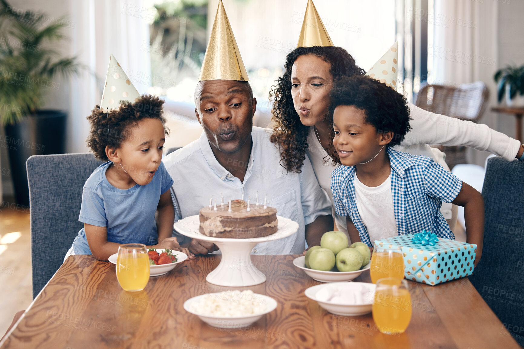 Buy stock photo Group of a young african american family wearing party hats and celebrating a birthday at home while blowing out candles on chocolate cake. Two adorable little boys with their parents having dessert
