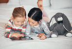 Two caucasian girls using a digital tablet while lying together on a bed at home. Happy young children browsing the internet online to play games and learn from educational apps before bedtime