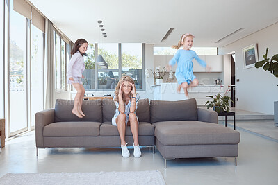 Fullbody caucasian woman sitting on a sofa at home and suffering from a headache while her hyperactive daughters jump around. Two adhd children annoying their mother by being naughty and misbehaving