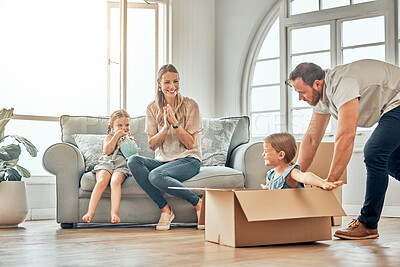 A happy mature father pushing his daughter in a box with mom and sister sit on the sofa at home. Man and girl having fun, playing games, while enjoying family bonding. Caucasian family in a new home