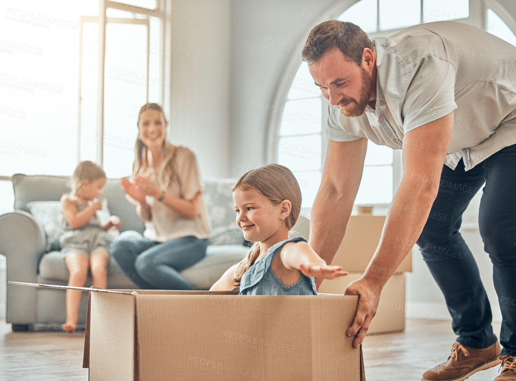 Buy stock photo A happy mature caucasian father pushing his daughter in a box while her mother and sister sit on the sofa in the lounge at home. Man and girl having fun, playing games at home while enjoying family bonding. Family looking overjoyed in their new home