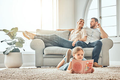 Buy stock photo Little girl reading storybook while lying on carpet at home. Smart kid reading book while relaxing at home with parents sitting on couch in background.