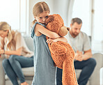 An upset little girl squeezing her teddy bear while looking sad and depressed while her parents argue in the background. Thinking about her parents breaking up or getting divorced is causing stress