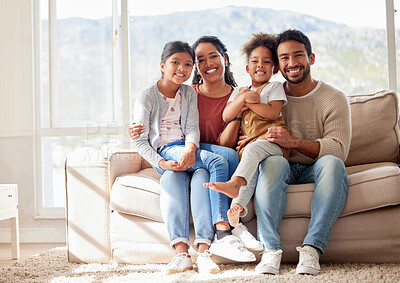 Portrait of two happy parents bonding with their daughters in the lounge, sitting on the sofa. Smiling young family relaxing at home, enjoying time together. Mother and father holding their daughters