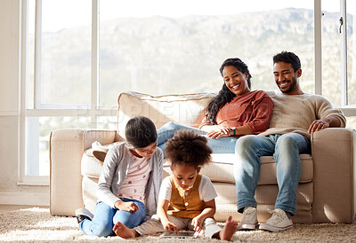 Buy stock photo Fullbody of a smiling young mixed race couple sitting close together on the sofa at home and watching their adorable children play. Cute hispanic girls bonding their mother and father in a living room