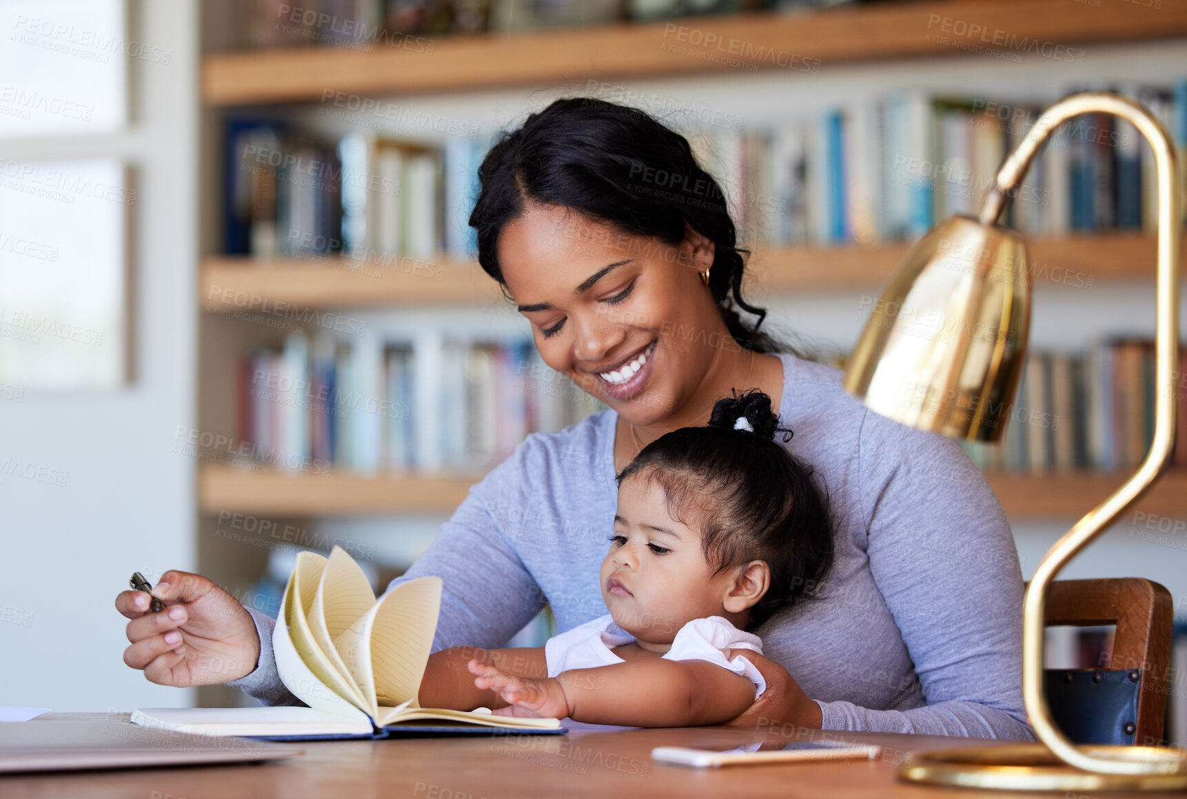 Buy stock photo Cheerful Mixed race mother sitting and smiling at a desk writing in a book with her baby daughter at home