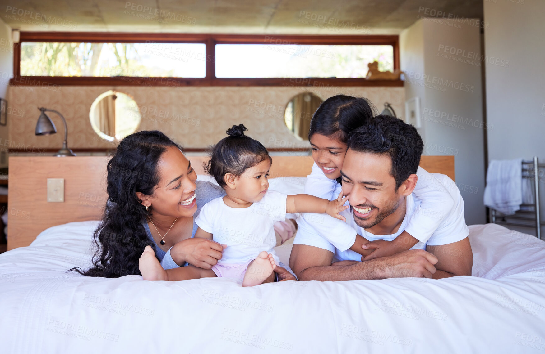 Buy stock photo Happy young family of four smiling and having fun in bed together at home. Young mixed race couple bonding with their two daughters and laughing together while looking comfortable in bed