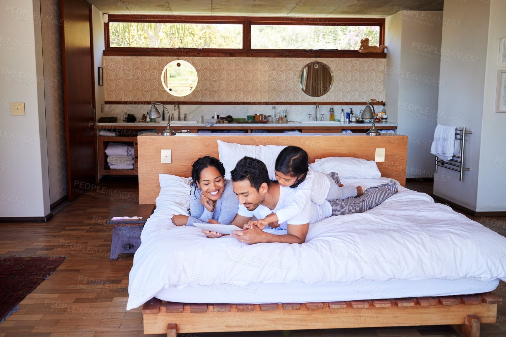 Buy stock photo A young mixed race family smiling and relaxing on the bed at home using a digital tablet.Two hispanic parents and their cute daughter watching videos on a wireless device while lying on the bed at home