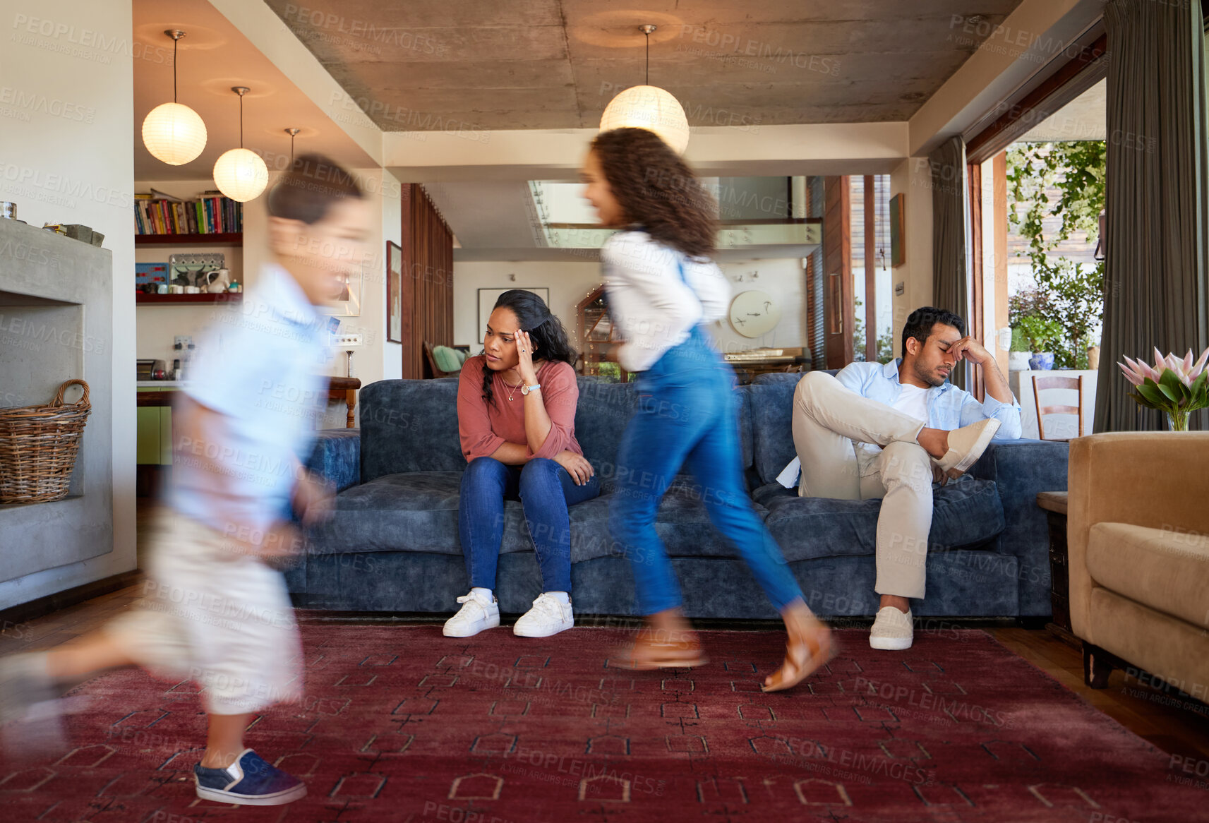 Buy stock photo Unhappy mixed race couple sitting on the sofa and looking stressed and miserable while their two young children with high energy are playfully running around them at home 