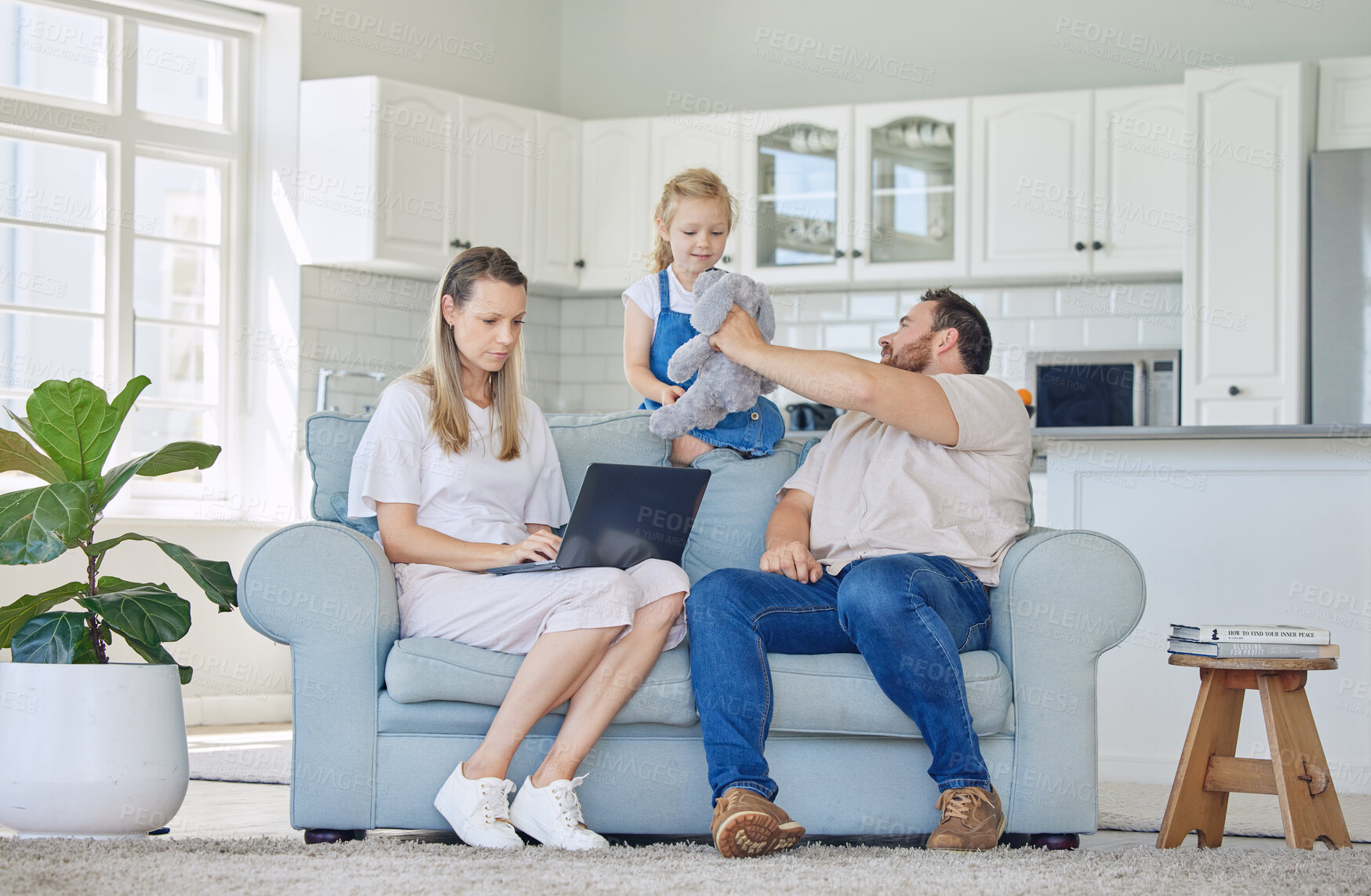 Buy stock photo Caucasian mother working on a laptop and sitting on the couch while her daughter and husband play with a teddybear in the lounge at home. Woman using a laptop. Father and child playing with a toy.