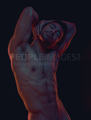 Sensual naked muscular man posing against dark studio background. Strong, nude man posing with his hands behind his head. Alluring man with six pack and muscles showing off his body. The male physique