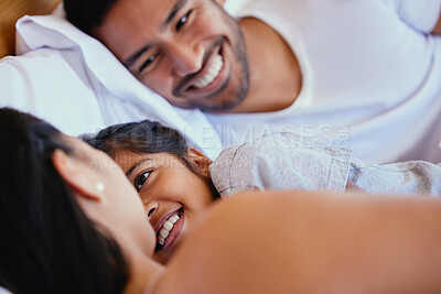 Buy stock photo Adorable little girl smiling while lying in bed with her parents. Mixed race family with one child relaxing and bonding in the morning while still lying in bedroom