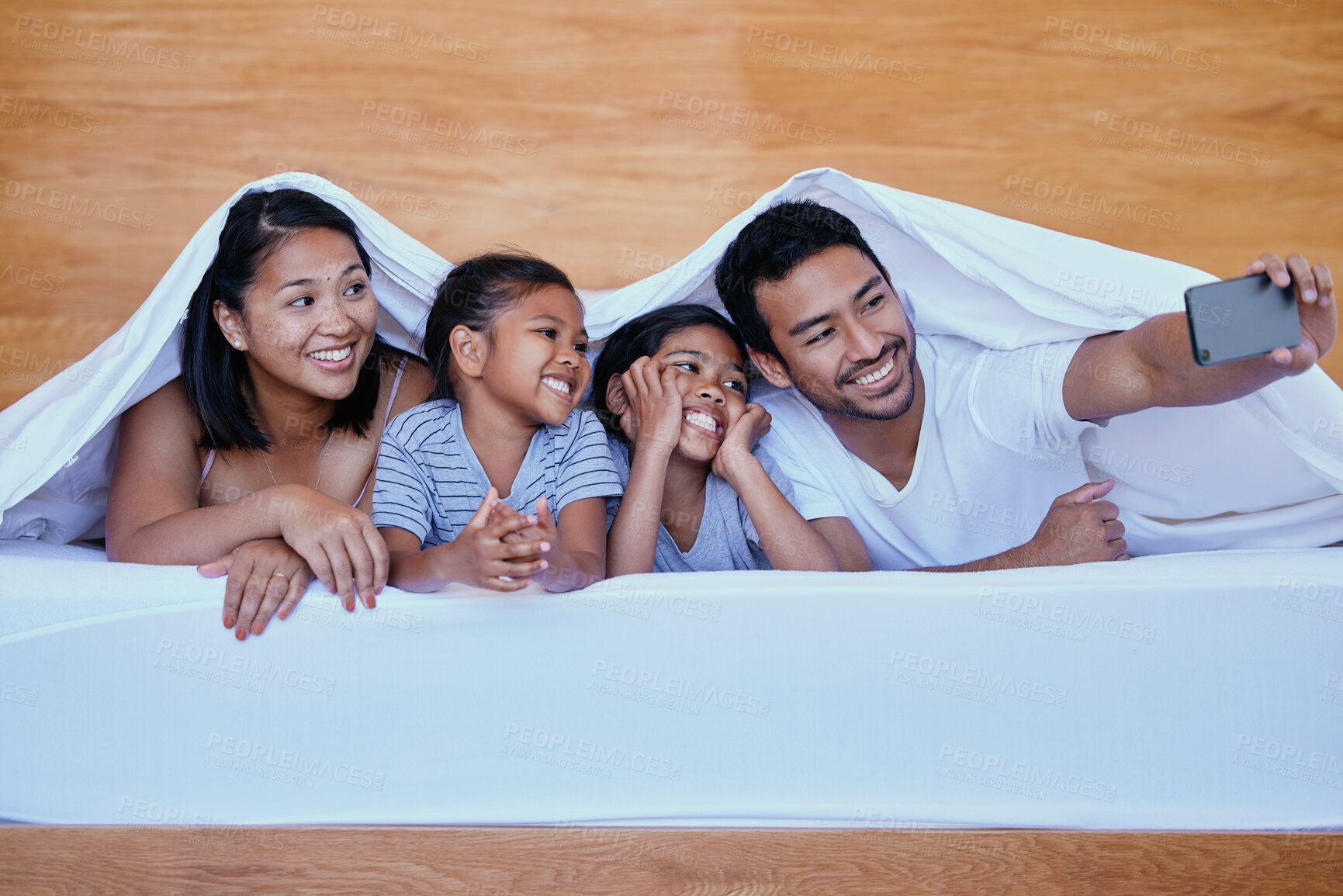Buy stock photo Happy family in bed. A young hispanic family in bed taking a selfie using a smartphone. Two parents bonding with their daughters at home. Sisters relaxing with their mother and father under bed sheets