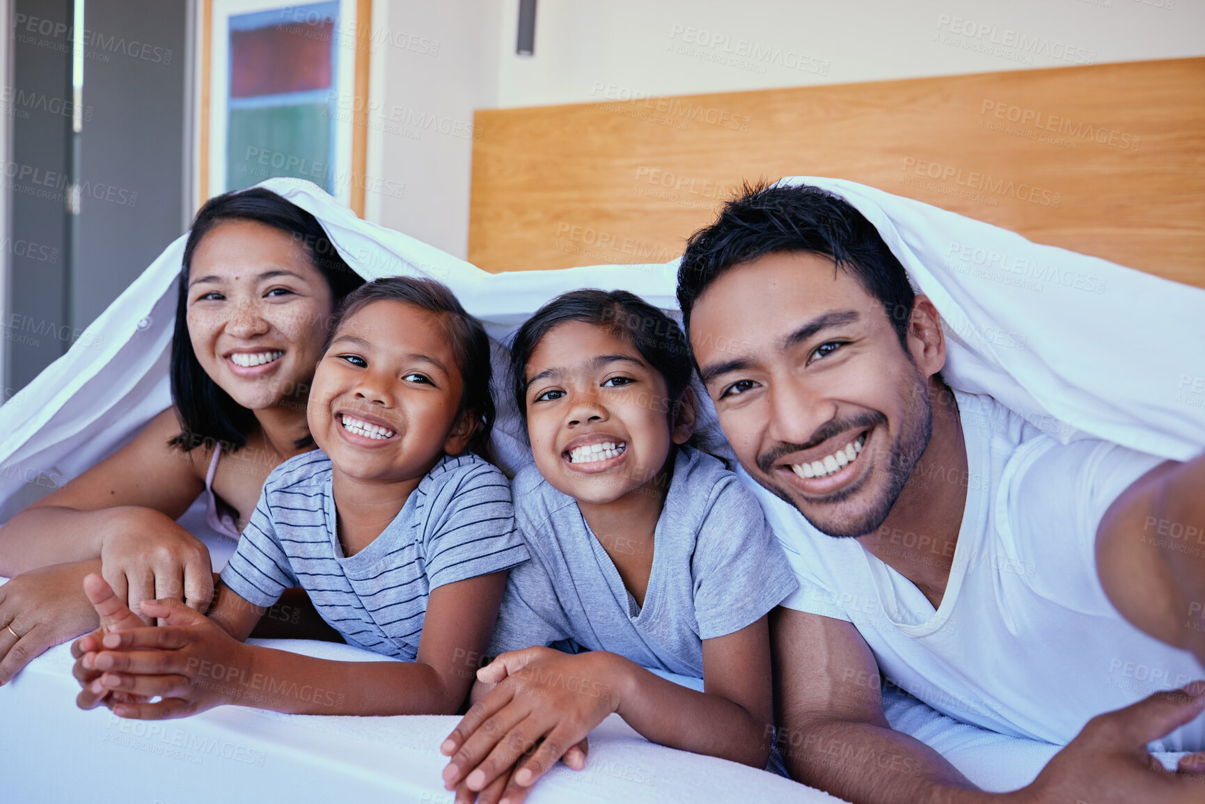 Buy stock photo Happy mixed race family of four in pyjamas lying cosy together in a row on a bed with blanket over their heads at home. Loving parents with two kids. Adorable girls bonding with mom and dad at bedtime
