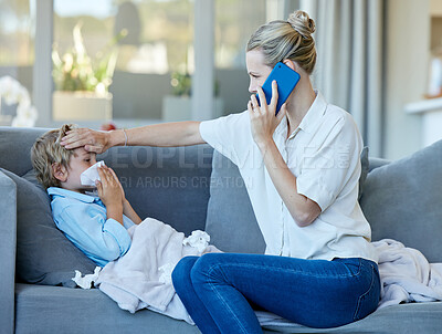 Young caucasian mother feeling her sick son\'s forehead while on a call using a phone sitting on the couch in the lounge at home. Ill child blowing his nose while sitting on the couch with his mom