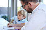 Young homeschool little boy sitting with his father at the kitchen table. Caucasian male single parent checking his son's homework and grading his paper at the kitchen table in a bright room 