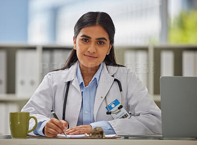 Portrait of a young indian doctor wearing a stethoscope sitting in a office writing a prescription while sitting at her desk. Mixed race Gp using a laptop and smiling