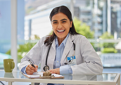 Portrait of happy indian doctor wearing a stethoscope sitting in a office writing a prescription at her desk. Mixed race Gp looking positive while doing paperwork against bright copyspace background