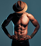 Fashionable african American model posing shirtless against blue studio background with copyspace. Sexy, unknown black man with attitude showing bare six pack while wearing hat. Masculine and muscle