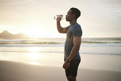 Fit man drinking water, taking a break. Young african american athlete exercising on the beach. Serious focused man drinking from a bottle. Confident black man resting during his workout outside