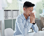 A sick businessman using a tissue and blowing his nose while working in a modern office. An ill asian man ill with flu, a cold or covid. Male employee sitting at a table and sneezing, feeling unhappy
