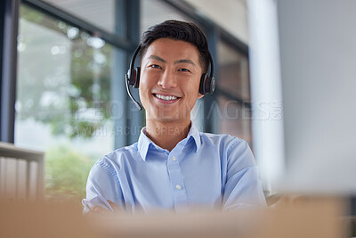Portrait of a professional asian man smiling and wearing a headset while sitting at his desk in an office job. Happy Asian call centre agent sitting at his desk in an office