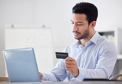 Young serious handsome mixed race businessman holding and using a credit card and working on a laptop in an office alone at work. Hispanic male boss making an online purchase on a laptop using a card