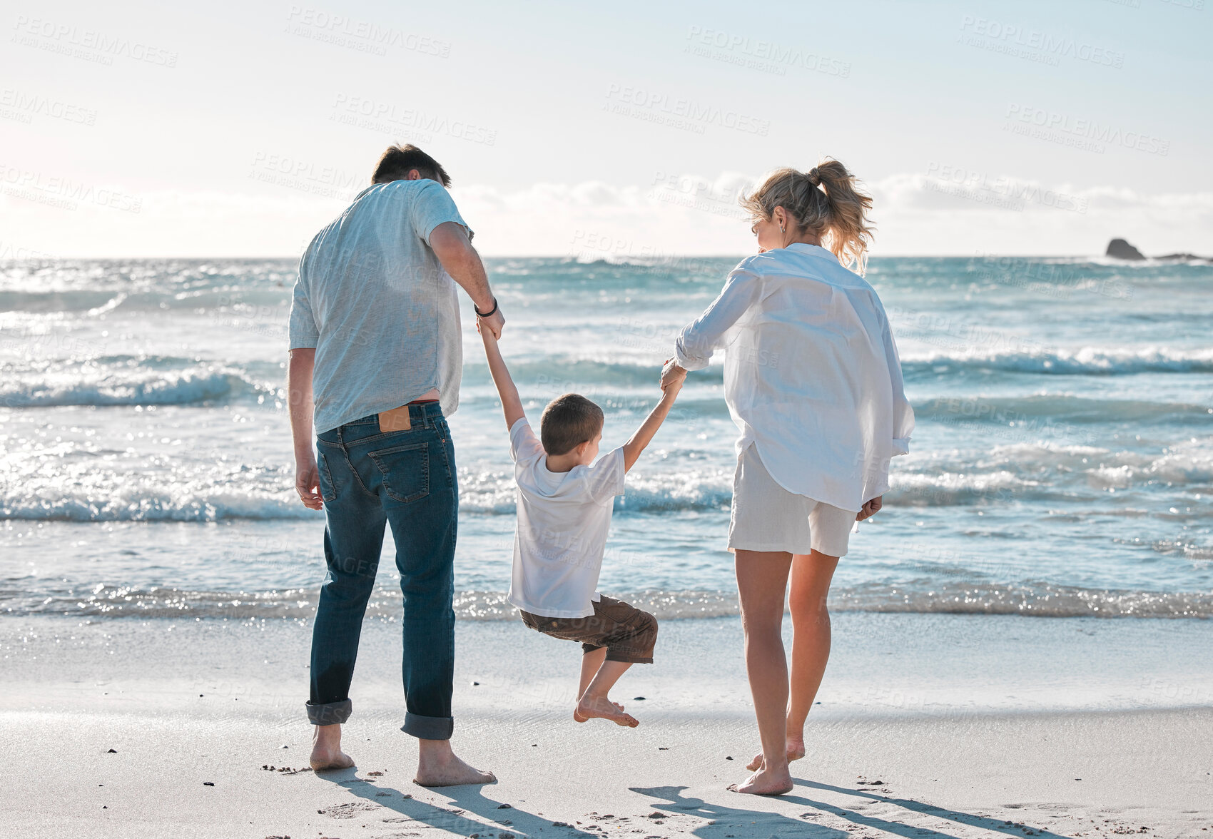 Buy stock photo Caucasian family from behind sharing quality time while enjoying a fun summer vacation at the beach. Loving parents swinging their playful little son by the arms while bonding and walking on the shore