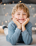 Portrait of one smiling little blonde caucasian boy lying on floor with his head resting in his hands at home. Face of adorable cheerful kid enjoying happy childhood with fairy lights in background