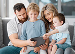 Happy relaxed family of four using a digital tablet device while bonding on the sofa together at home. Young couple checking social media and watching a movie with their two cute caucasian sons 