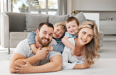 Portrait of smiling caucasian family of four relaxing on floor in lounge at home. Playful sons lying and clinging on carefree loving parents\' backs while bonding and spending fun quality time together