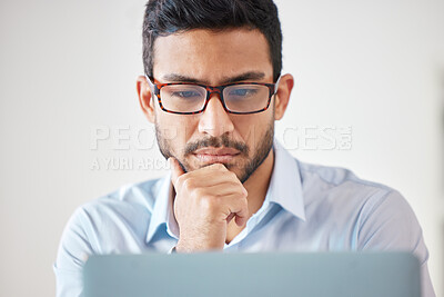 One focused mixed race businessman wearing glasses thinking of ideas while planning to brainstorm on a laptop in an office. Young motivated entrepreneur searching for inspiration and making decisions