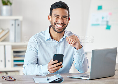 Portrait of a young handsome happy mixed race businessman using social media on his phone while working alone on a laptop in an office at work. One hispanic male boss smiling using a cellphone at work