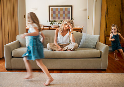 Young unhappy caucasian mother suffering from a headache with her daughters running and playing in the lounge at home. Little siblings being playful together while their mom is stressed sitting on the couch. Woman upset while her children play