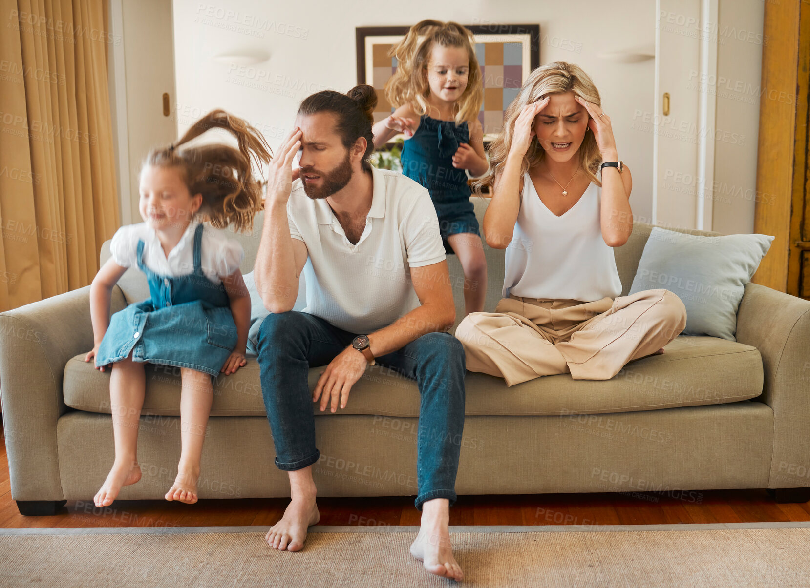 Buy stock photo Young caucasian mother and father suffering a headache with their daughters playing jumping on the couch in the lounge at home. Little sisters being playful while their parents are stressed. Woman and man tired sitting on the couch